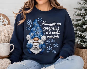 May include: A navy blue crewneck sweatshirt featuring a graphic of a gnome wearing a blue hat with white snowflakes. The gnome holds a mug labeled HOT COCOA. The text reads: Snuggle up gnomies it's cold outside, surrounded by white snowflakes.