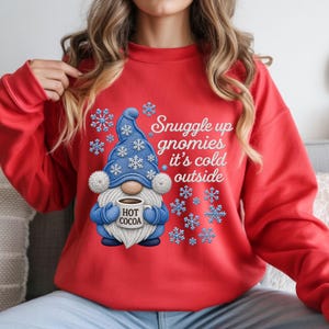May include: A bright red long-sleeve sweatshirt featuring a graphic of a blue gnome wearing a hat with white snowflakes. The gnome holds a mug labeled HOT COCOA. The text reads Snuggle up gnomies it's cold outside, surrounded by blue snowflakes.