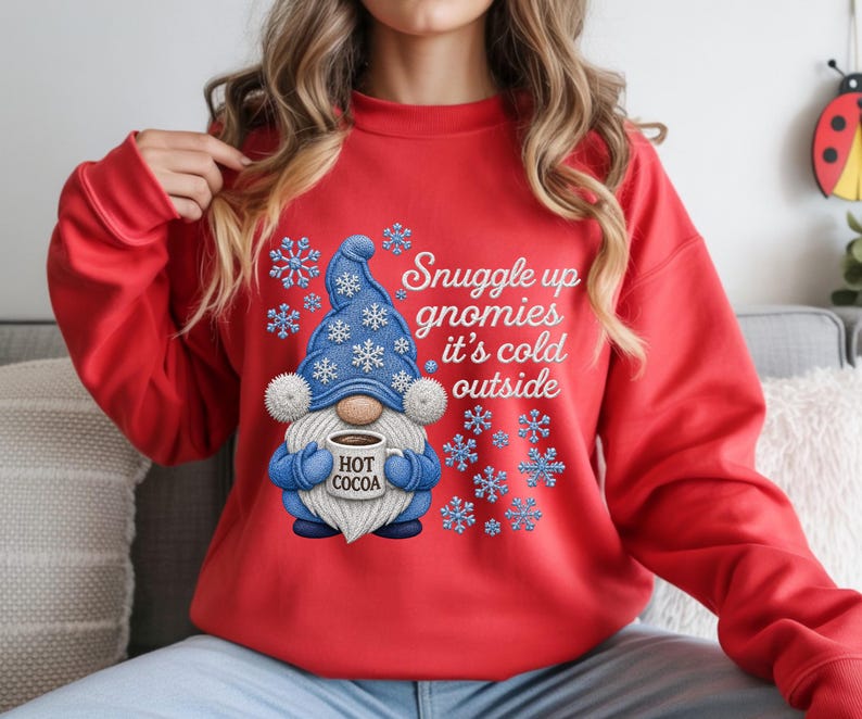 May include: A bright red long-sleeve sweatshirt featuring a graphic of a blue gnome wearing a hat with white snowflakes. The gnome holds a mug labeled HOT COCOA. The text reads Snuggle up gnomies it's cold outside, surrounded by blue snowflakes.