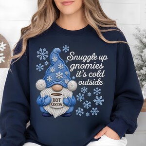 May include: A navy blue crewneck sweatshirt featuring a graphic of a gnome wearing a blue hat with white snowflakes. The gnome holds a mug labeled HOT COCOA. The text reads Snuggle up gnomies it's cold outside, surrounded by blue and white snowflakes.