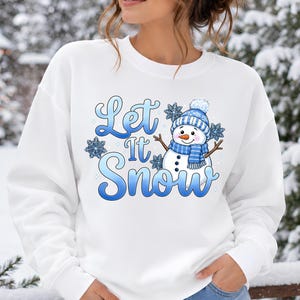 May include: A white crewneck sweatshirt featuring a graphic print of a smiling snowman wearing a blue striped hat and scarf. The text Let It Snow is written in blue gradient lettering, surrounded by several blue and silver snowflake illustrations.