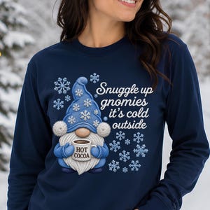 May include: A navy blue long-sleeve sweatshirt featuring a graphic of a blue gnome wearing a matching hat with white snowflakes. The gnome holds a white mug labeled HOT COCOA. The text reads Snuggle up gnomies it's cold outside.