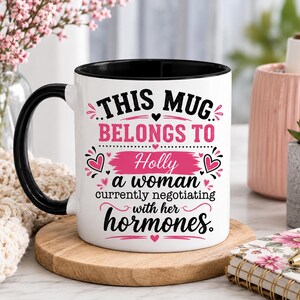 May include: A white ceramic mug with a black handle and rim, featuring pink and black text that reads: This mug belongs to Holly a woman currently negotiating with her hormones. The design includes small heart icons and decorative flourishes.