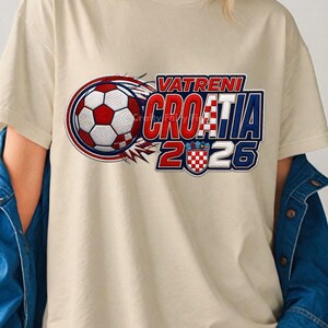 May include: Cream-colored t-shirt featuring a soccer ball graphic with red, white, and blue accents. The text "VATRENI CROATIA 2026" is displayed in a bold, stylized font, with the Croatian flag incorporated into the design.