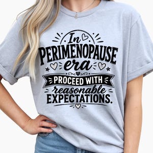 May include: A light blue short-sleeve t-shirt featuring black text that reads In PERIMENOPAUSE era PROCEED WITH reasonable EXPECTATIONS. The design includes small heart and star graphics surrounding the lettering, printed on the front of the shirt.