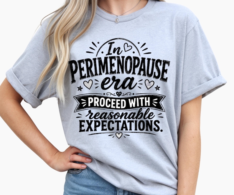 May include: A light blue short-sleeve t-shirt featuring black text that reads In PERIMENOPAUSE era PROCEED WITH reasonable EXPECTATIONS. The design includes small heart and star graphics surrounding the lettering, printed on the front of the shirt.