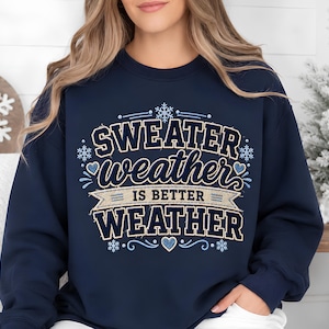 May include: A navy blue crewneck sweatshirt featuring a graphic design with the text "SWEATER weather IS BETTER WEATHER" in white and light blue lettering, accented with small snowflakes and heart shapes in a decorative, layered style.