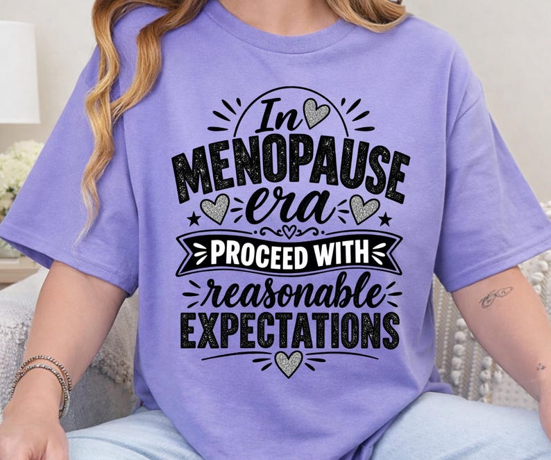 May include: A light purple short-sleeved t-shirt featuring black text that reads In MENOPAUSE era PROCEED WITH reasonable EXPECTATIONS. The design includes decorative hearts and stars, with the word era in a script font and other words in block lettering.