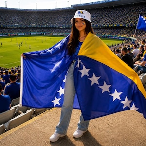 May include: A person wearing a white bucket hat with the text "BOSNIA" and a blue and yellow flag with white stars. The person is standing in a stadium with a soccer field in the background.