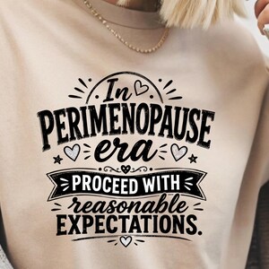 May include: A beige t-shirt featuring black text that reads In PERIMENOPAUSE era PROCEED WITH reasonable EXPECTATIONS. The design includes small heart and star icons, with a decorative banner element surrounding the middle text lines.
