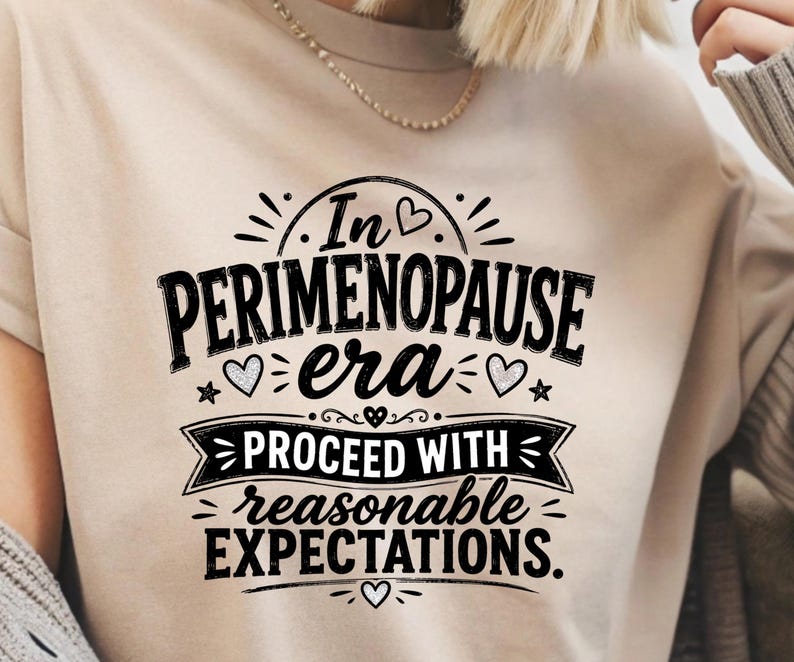May include: A beige t-shirt featuring black text that reads In PERIMENOPAUSE era PROCEED WITH reasonable EXPECTATIONS. The design includes small heart and star icons, with a decorative banner element surrounding the middle text lines.
