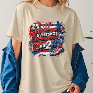 May include: Beige t-shirt featuring a colorful graphic design with the text "HRVATSKA SVJETSKO PRVENSTVO 2026". The design includes a soccer ball, a soccer player, a soccer shoe, and the Croatian flag colors.