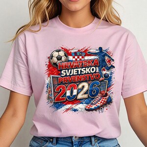 May include: A light pink t-shirt featuring a soccer-themed graphic. The design includes a soccer ball, a soccer player, and the text "HRVATSKA SVJETSKO PRVENSTVO 2026" in red, white, and blue, with a Croatian flag.