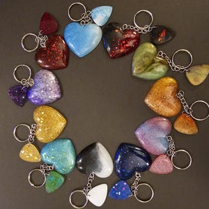 May include: A collection of 18 heart-shaped keychains with various colors and glitter accents. The keychains are arranged in a circle on a dark surface.