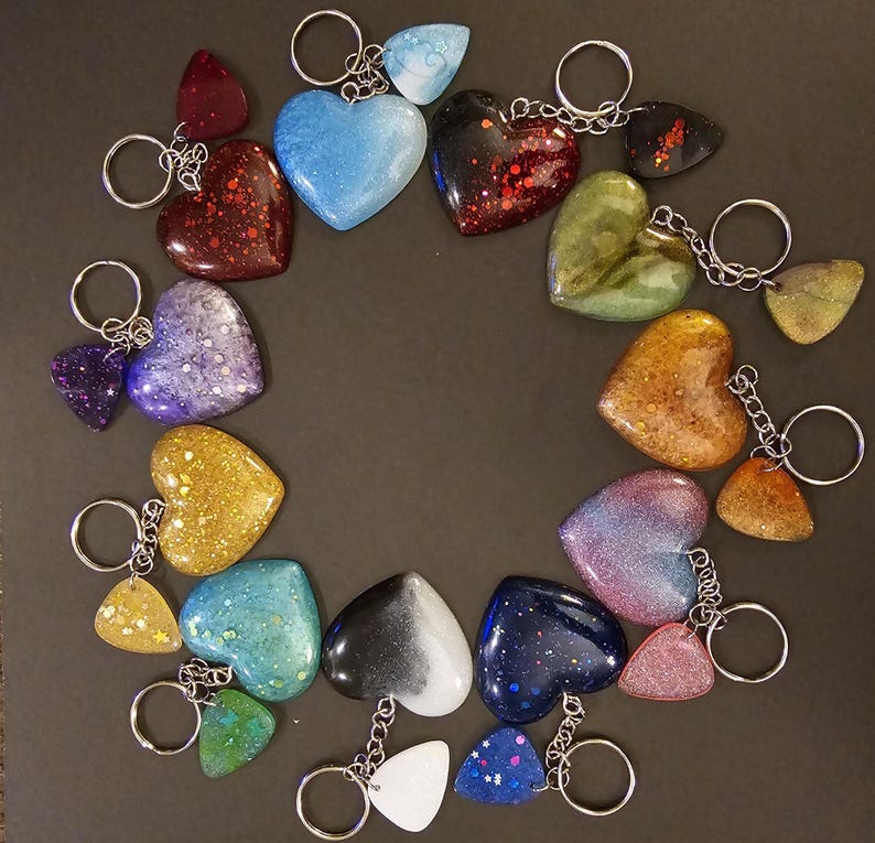 May include: A collection of 18 heart-shaped keychains with various colors and glitter accents. The keychains are arranged in a circle on a dark surface.