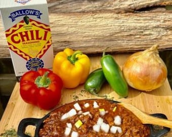 May include: A bowl of chili with onions and peppers. The chili is topped with chopped onions and a few yellow peppers. The bowl is sitting on a wooden surface. There is a box of chili seasoning in the background.