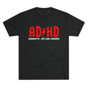 Puede incluir: Camiseta gris oscuro con el texto rojo "ADHD" y un rayo. Debajo, el texto blanco dice "HIGHWAY TO... HEY LOOK, DRAGONS". La camiseta tiene cuello redondo y mangas cortas.