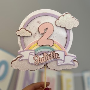Peut inclure: Un topper de gâteau rose et blanc avec un arc-en-ciel, des nuages et le chiffre 2. Le topper a également une bannière avec le nom "Juliette" écrit en rose.