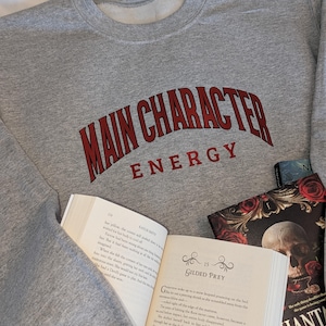 Peut inclure: Un sweat-shirt gris avec l'inscription "MAIN CHARACTER ENERGY" en lettres rouges arquées. Un livre ouvert et deux autres livres sont visibles, suggérant un thème de lecture.