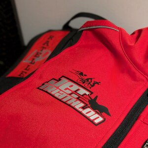May include: Red jacket with a black zip and black trim. The jacket has a silver logo that says "Jet Triathlon" with a silhouette of a person running, cycling, and swimming.