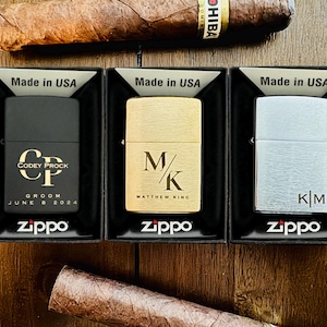 May include: Three silver, gold and black Zippo lighters with personalised engravings. The lighters are in their boxes, which are black with white text that reads "Made in USA".