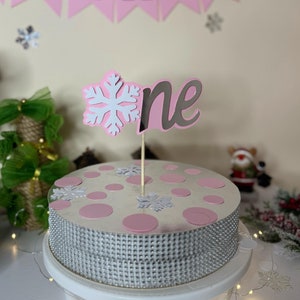 May include: A white cake with pink polka dots and a silver glitter border. The cake is decorated with a white snowflake and a pink "one" topper. The cake is on a white cake stand with a string of white lights.