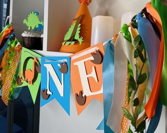 Könnte beinhalten: Eine farbenfrohe Dinosaurier-Geburtstagsbanner mit dem Wort "ONE" auf einzelnen Wimpeln. Das Banner zeigt grüne, blaue und orangefarbene Flaggen, Dinosaurier-Fußabdrücke und dekorative Bänder. Ein passender Partyhut und Cupcakes sind ebenfalls zu sehen.