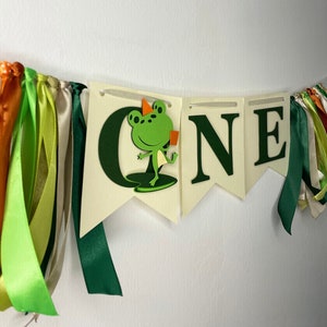 May include: A decorative banner spelling "ONE" in dark green letters. The letter "O" features a green frog with an orange party hat. The banner is adorned with colourful ribbons in shades of green, orange, and cream.