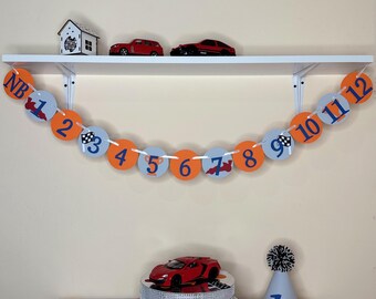 May include: A red race car cake topper on a white cake stand with a silver sparkly base. A white and gray party hat with a black pom-pom on top. A banner with the letters NB and numbers 1 through 12 in blue and orange circles. The banner is hanging on a white shelf with two red toy cars.