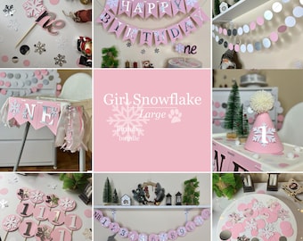 May include: Pink and silver snowflake themed first birthday party decorations. The decorations include a banner that says "Happy Birthday", a high chair banner that says "One", a number one cake topper, a snowflake hat, and a garland of pink and silver circles.