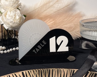 May include: Laser cut table number template in the shape of a heart. The number 12 is white on a black background. The left side of the heart is white with a sunburst design and the text "TABLE" in black.