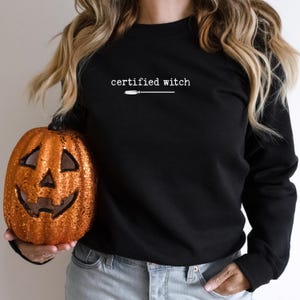 May include: Black sweatshirt with the text "certified witch" in white, along with a broom graphic. A person is holding a glittery orange pumpkin with a carved jack-o'-lantern face. The sweatshirt and pumpkin are Halloween-themed.