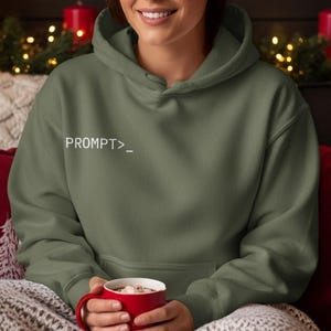 Peut inclure: Un sweat à capuche vert olive avec le texte blanc "PROMPT>_". Le sweat-shirt à capuche a une poche avant et un cordon de serrage. La personne tient une tasse rouge.