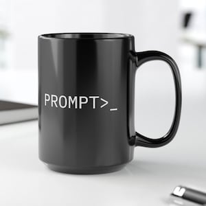 May include: A black ceramic coffee mug with a large handle. The mug features the white text "PROMPT>_". The mug is sitting on a white surface.