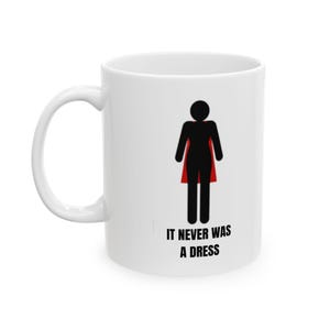 Könnte beinhalten: Weiße Keramiktasse mit einer schwarzen Silhouette einer Person, die einen roten Umhang trägt. Der Text "IT NEVER WAS A DRESS" ist unter dem Bild aufgedruckt.