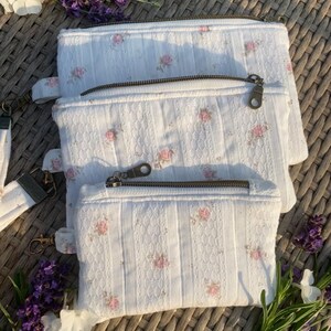 Puede incluir: Tres estuches blancos con cremallera de diferentes tamaños, confeccionados en tela texturizada con un delicado estampado floral rosa. Cada estuche rectangular incluye un cierre de cremallera metálico y una pequeña trabilla de tela lateral.