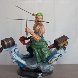 May include: A green and brown figurine of a character from the anime One Piece. The character is holding two swords and has a green bandana around his head. The figurine is standing on a base with a blue and white wave design. There is a barrel and a treasure chest on the base.