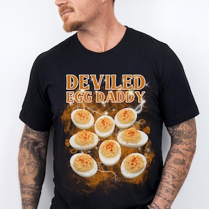 May include: Black t-shirt with the text "DEVILED EGG DADDY" in orange and white. The shirt features a graphic of deviled eggs with paprika. The shirt is a casual wear item.