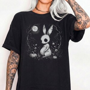 May include: A black t-shirt featuring a white graphic illustration of a rabbit sitting in a field with pumpkins under a full moon. The design includes intricate line work depicting flowers and swirling branches surrounding the central rabbit figure.