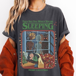 Op de afbeelding: Donkergrijs t-shirt met de tekst "THEY SEE YOU WHEN YOU'RE SLEEPING" boven een raamsc&egrave;ne met monsters en een meisje in bed. Het shirt zegt ook "MERRY CREEPMAS".