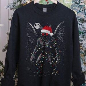 K&ouml;nnte beinhalten: Schwarzes Sweatshirt mit einer Grafik einer mothman-&auml;hnlichen Kreatur, die einen Weihnachtsmannhut und Lichterketten tr&auml;gt. Das Design beinhaltet einen Vollmond und eine Silhouette des Weihnachtsmanns. Das Sweatshirt wird auf einem Holzb&uuml;gel pr&auml;sentiert.
