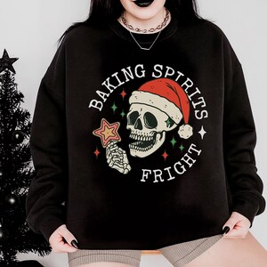 Könnte beinhalten: Schwarzes Sweatshirt mit einem Grafikdesign, das einen Totenkopf mit Weihnachtsmütze und einem sternförmigen Keks zeigt. Der Text lautet "BAKING SPIRITS FRIGHT" in einem kreisförmigen Design. Ein kleiner schwarzer Weihnachtsbaum ist im Hintergrund.