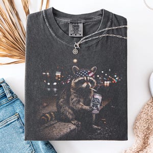 K&ouml;nnte beinhalten: Dunkelgraues Comfort Colors T-Shirt mit einem Waschb&auml;ren, der ein Bandana mit amerikanischer Flagge tr&auml;gt, eine Zigarette raucht und eine Bierdose h&auml;lt. Das T-Shirt hat einen Rundhalsausschnitt und kurze &Auml;rmel. Eine silberne Halskette liegt &uuml;ber dem T-Shirt.