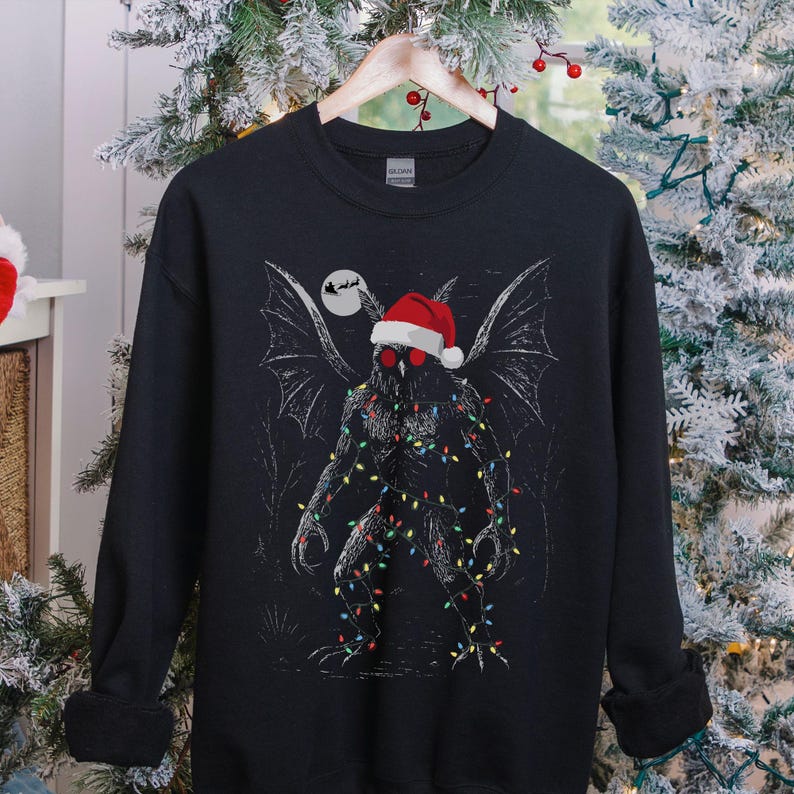 K&ouml;nnte beinhalten: Schwarzes Sweatshirt mit einer Grafik einer mothman-&auml;hnlichen Kreatur, die einen Weihnachtsmannhut und Lichterketten tr&auml;gt. Das Design beinhaltet einen Vollmond und eine Silhouette des Weihnachtsmanns. Das Sweatshirt wird auf einem Holzb&uuml;gel pr&auml;sentiert.