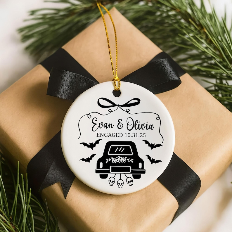 May include: White ceramic ornament with a gold string, featuring a black car illustration, bats, and the names "Evan & Olivia" with the date "ENGAGED 10.31.25". The ornament is on a wrapped gift with a black ribbon.