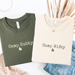 Könnte beinhalten: Zwei T-Shirts, eines in Olivgrün und eines in Creme, werden gezeigt. Das olivgrüne Shirt trägt den Aufdruck "Camp Hubby" mit einer kleinen Baumgrafik. Das cremefarbene Shirt trägt "Camp Wifey" mit einer ähnlichen Baumgrafik. Beide sind gefaltet.