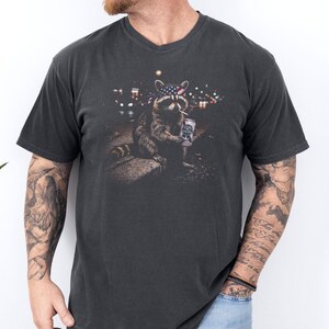 K&ouml;nnte beinhalten: Dunkelgraues V-Ausschnitt-T-Shirt mit einem Waschb&auml;ren-Motiv. Der Waschb&auml;r tr&auml;gt ein amerikanisches Flaggen-Bandana, raucht eine Zigarette und h&auml;lt eine Bierdose. Im Hintergrund ist eine Stadt bei Nacht zu sehen.
