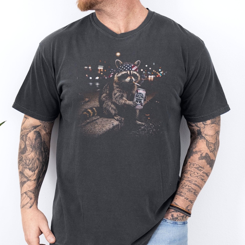 K&ouml;nnte beinhalten: Dunkelgraues V-Ausschnitt-T-Shirt mit einem Waschb&auml;ren-Motiv. Der Waschb&auml;r tr&auml;gt ein amerikanisches Flaggen-Bandana, raucht eine Zigarette und h&auml;lt eine Bierdose. Im Hintergrund ist eine Stadt bei Nacht zu sehen.