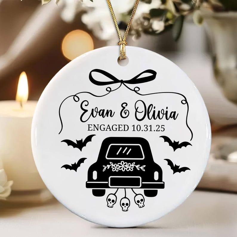 May include: White ceramic ornament with a gold string for hanging. The ornament features the names "Evan & Olivia" and the date "10.31.25" with a black car, bats, and skulls design. A decorative item for a wedding or engagement.
