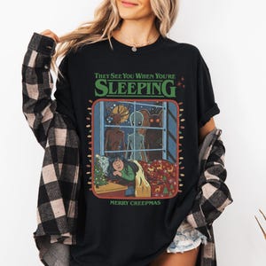 Op de afbeelding: Een zwart t-shirt met een grafische print met de tekst "THEY SEE YOU WHEN YOU'RE SLEEPING" en "MERRY CREEPMAS". De illustratie toont drie wezens die door een raam naar een persoon in bed kijken, omringd door kerstverlichting en decoraties.